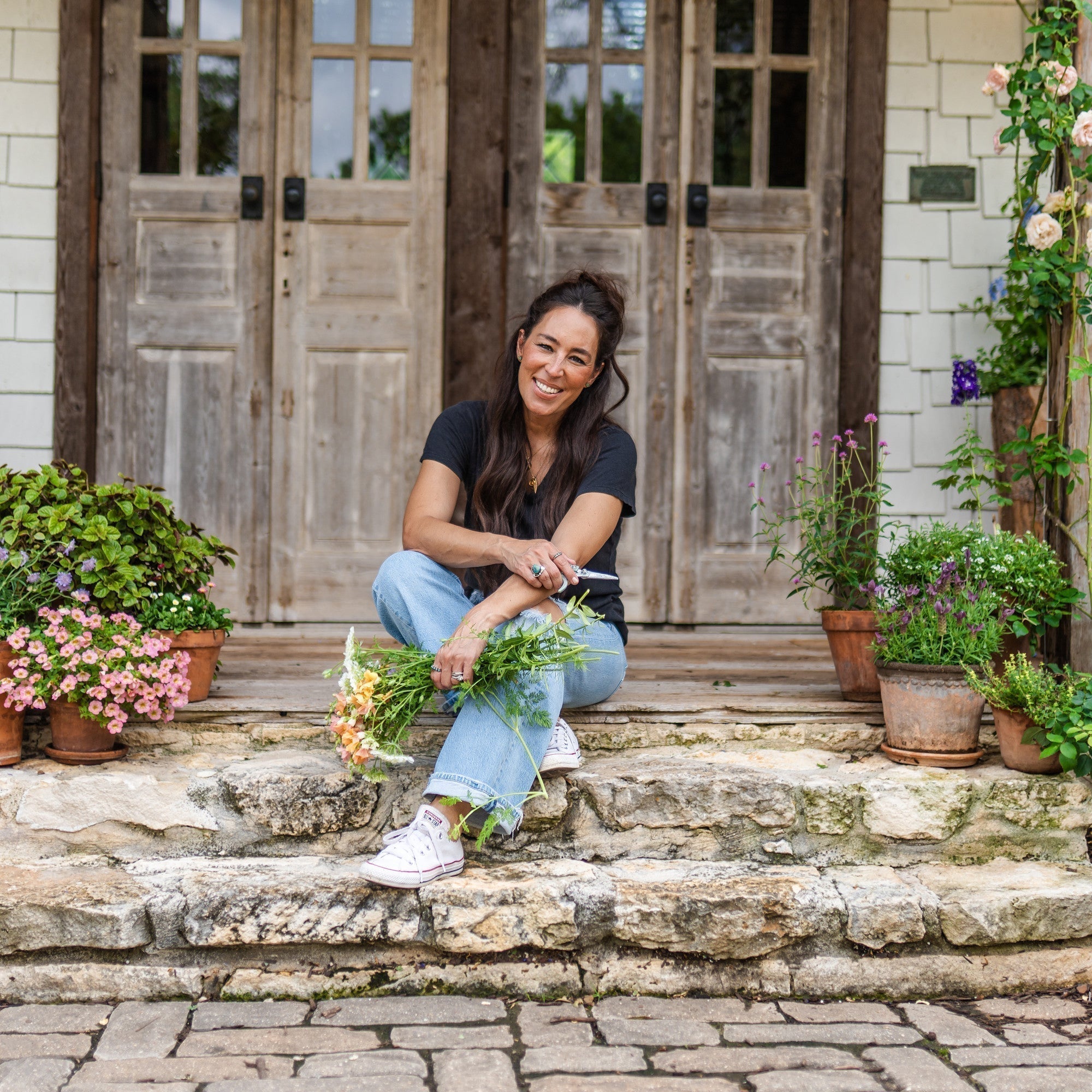 Joanna Gaines smiles on her front porch—surrounded by plants and holding a bundle of fresh flowers.
