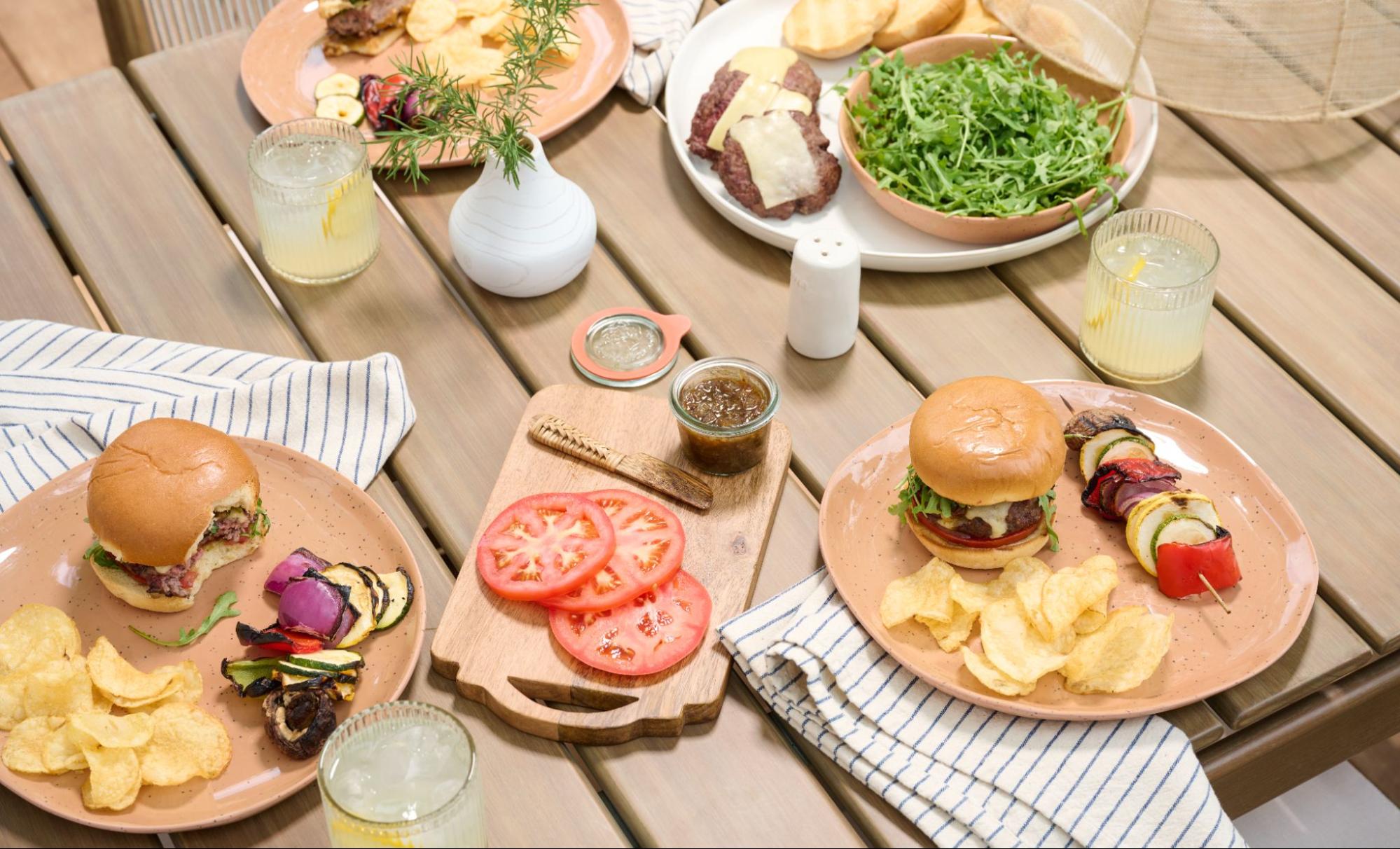 A summer picnic spread with burgers, potato chips, tomatoes, grilled skewers, and more. 