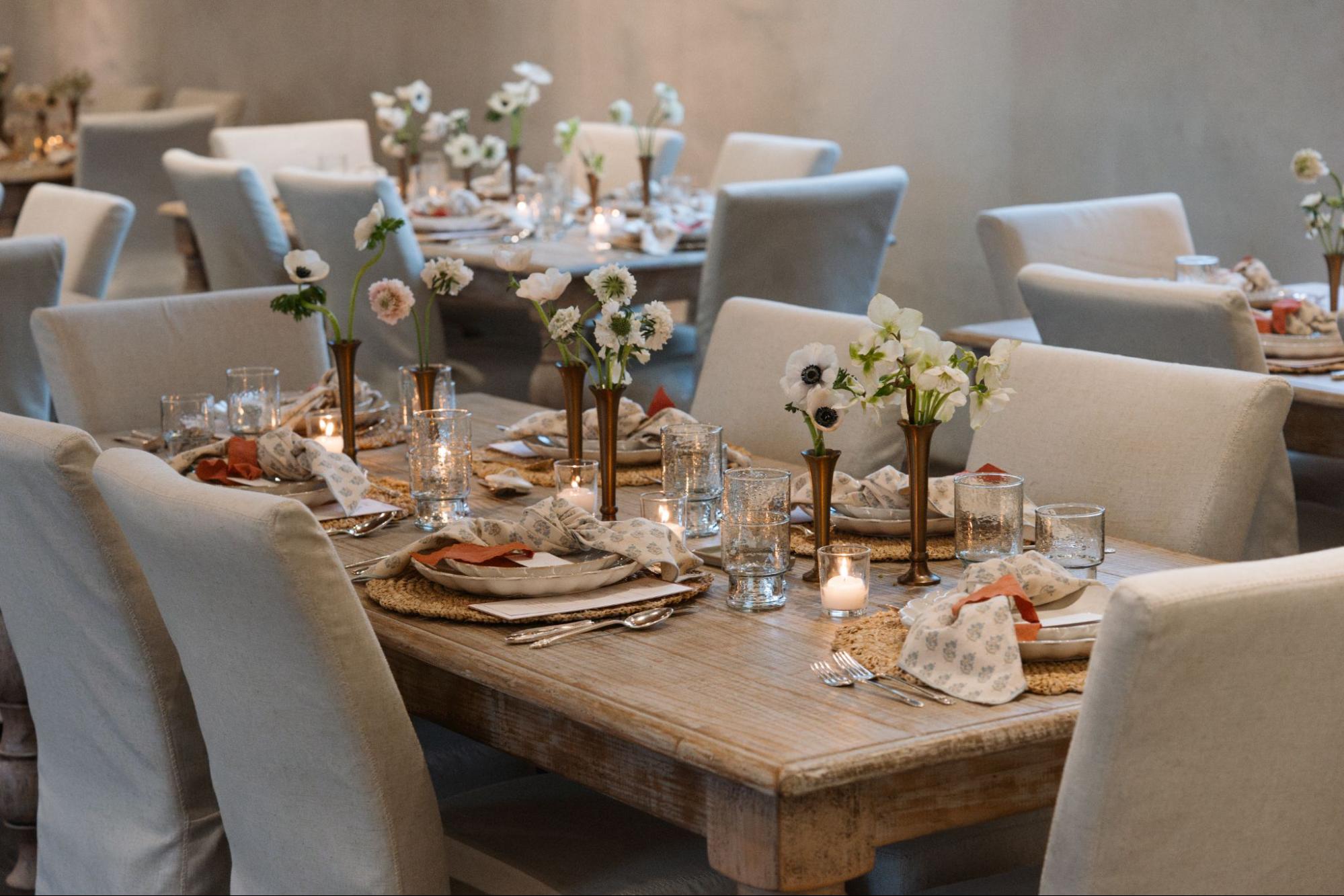 A wooden table from Magnoliass is topped with a spring-inspired tablescape, including block-printed napkins, brass bud vases, scalloped dishes, and more. 