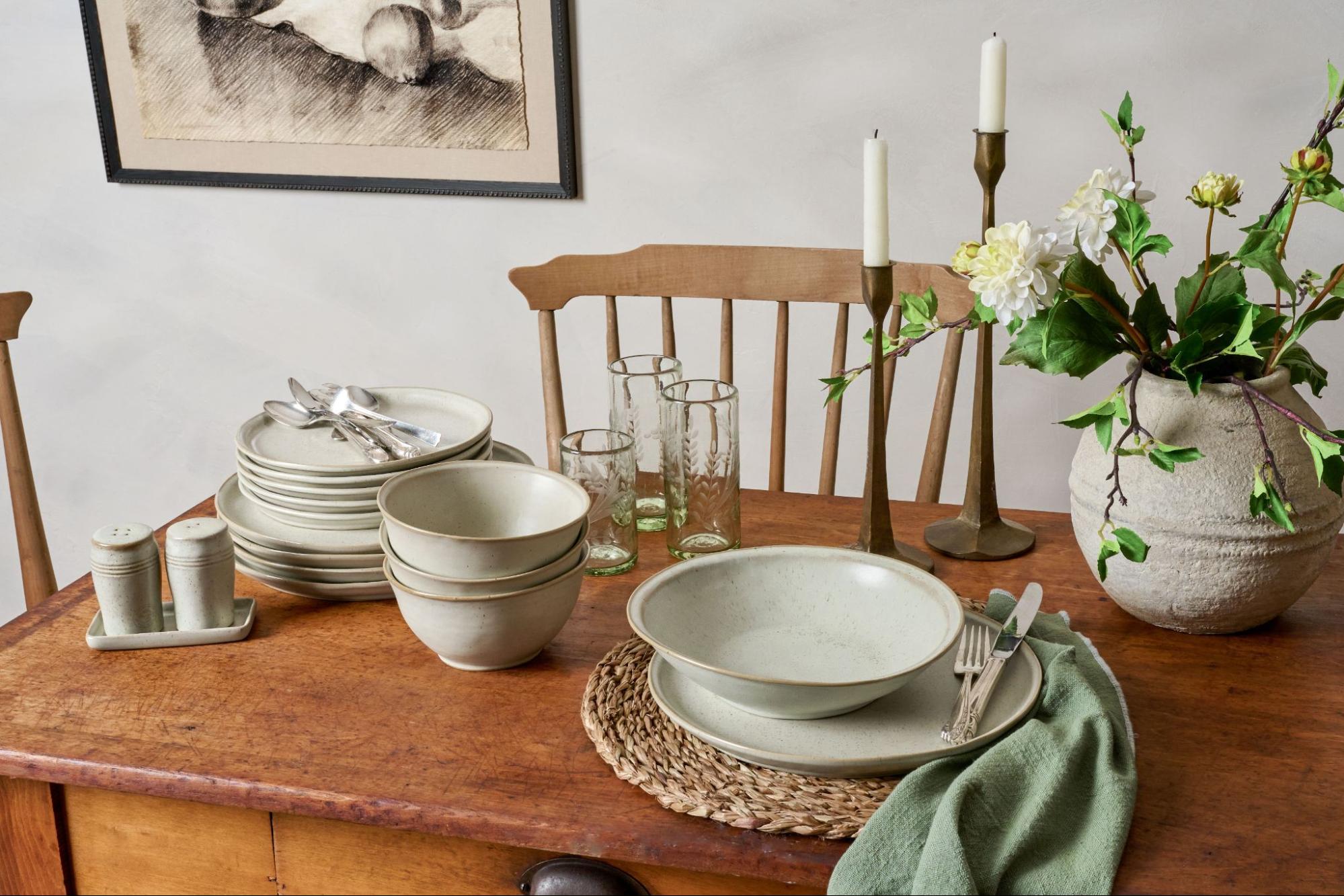 Magnoliass tableware sits on a wooden table next to taper candles and a grey vase with faux blooms.