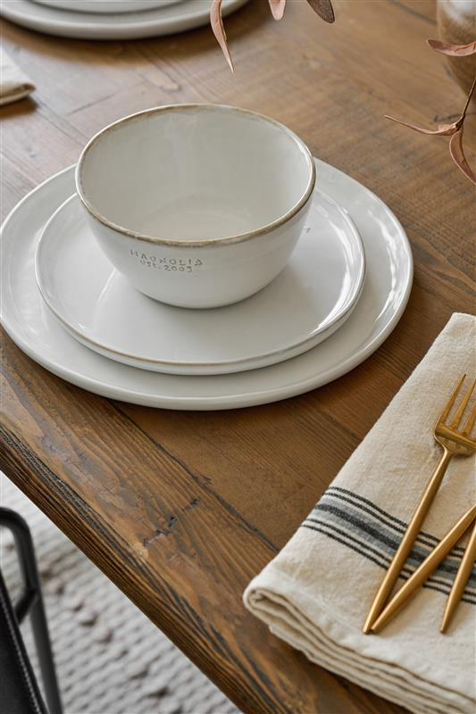 Two plates and a bowl from Magnoliass sit on a wooden table next to a striped napkin and gold flatware.