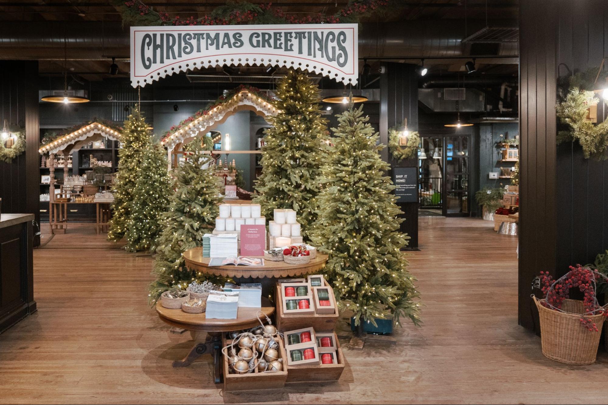 Faux evergreen trees surround a table with Magnoliass merchandise—topped by a sign that says "Christmas Greetings."