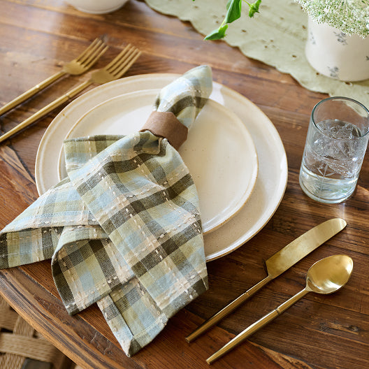 Set table with plaid napkins, gold cutlery, and a glass of water on a wooden surface.