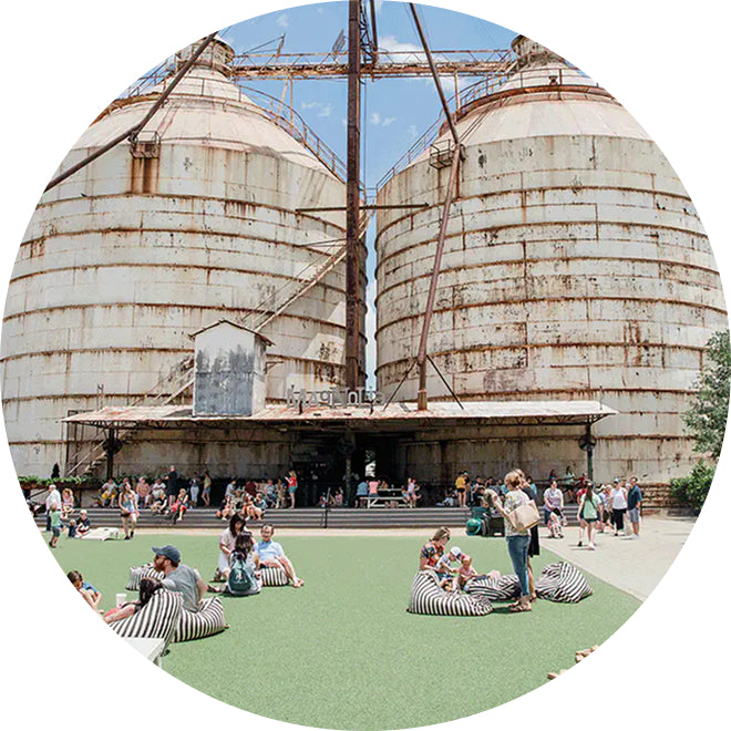 People sitting on grass in front of large rusted industrial tanks on the Silos grounds.