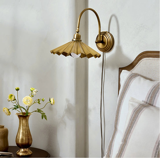 Gold wall sconce on a white wall next to a vase with flowers and a bed.