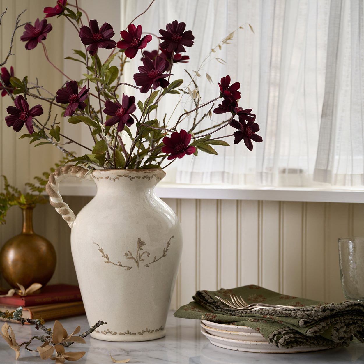 White vase with purple flowers on a table with a window in the background.  Shop Florals and Vases.