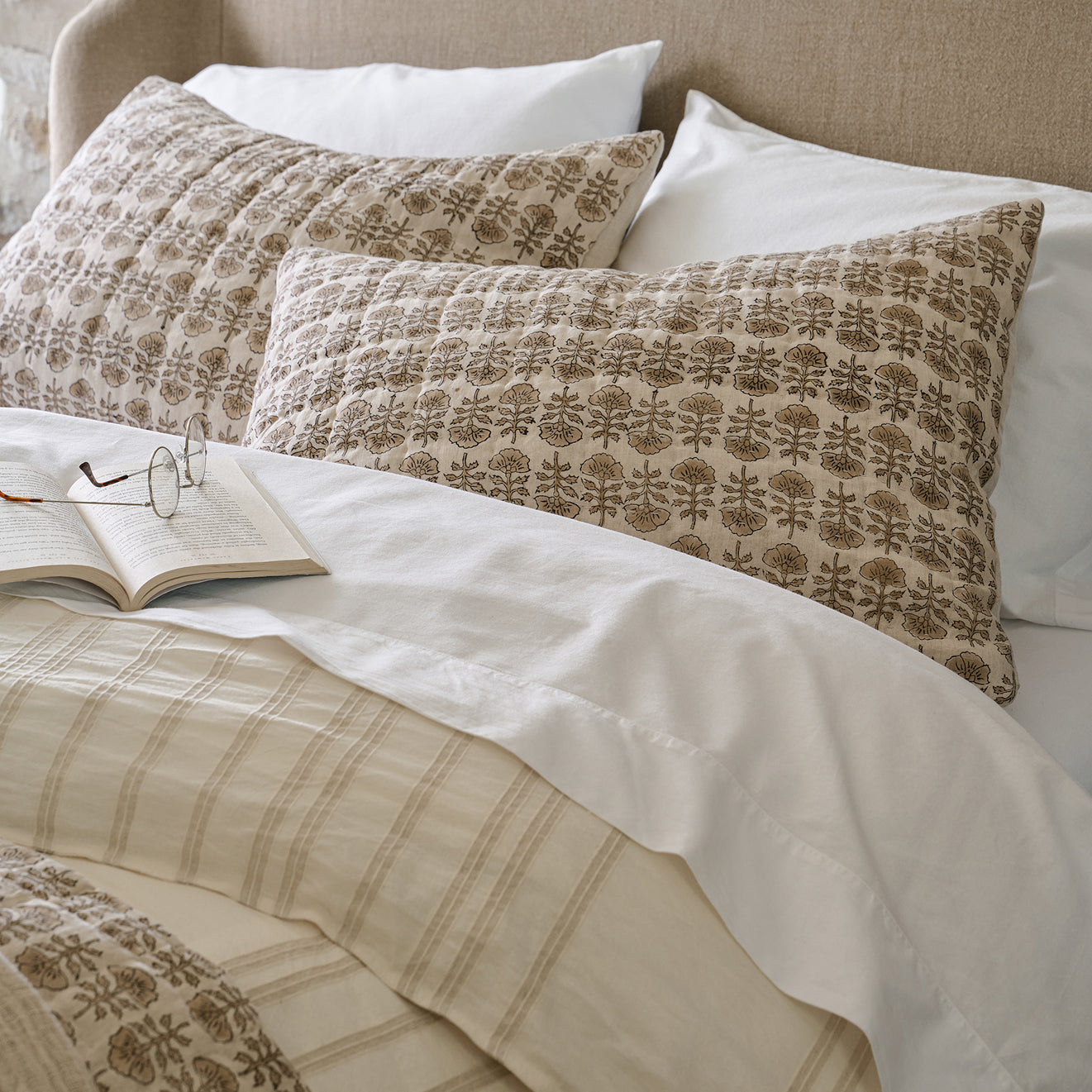Patterned pillows on a bed with an open book and glasses