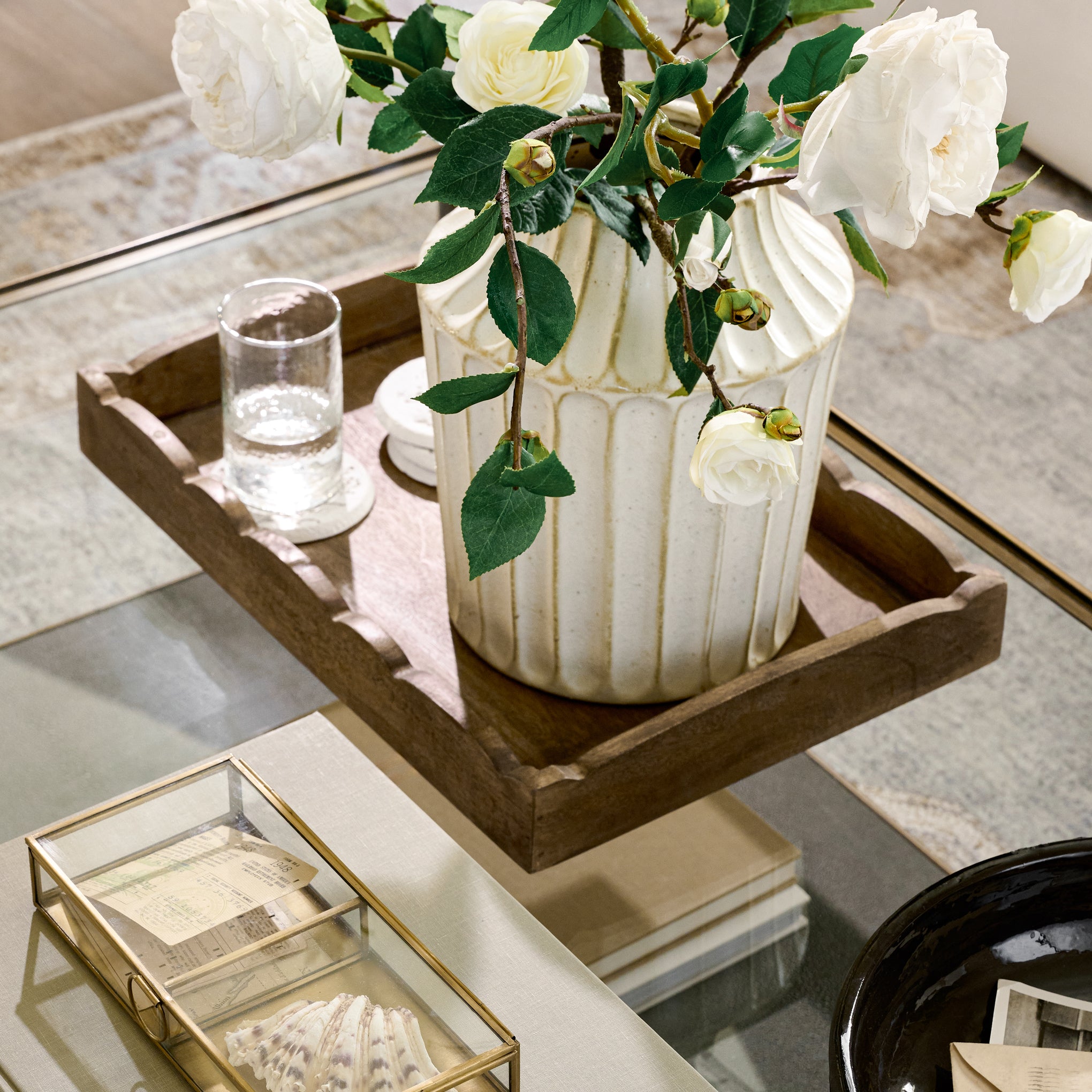 Scalloped Antiqued Wood Tray on a glass coffee table with a vase and a glass of water on the tray