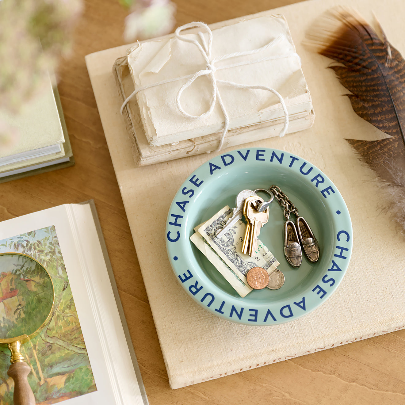 Small blue dish with 'Chase Adventure' text holding keys, coins, and a small shoe on a wooden surface with books and a feather.