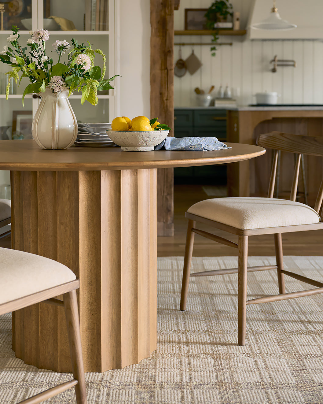 Dining room with wooden table and chairs, flowers, and kitchen in the background.