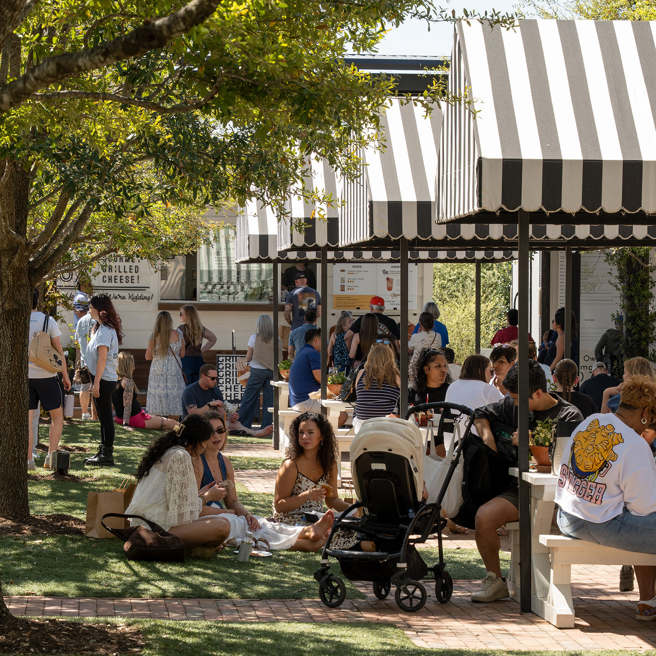 People sitting outdoors under a striped canopy with trees and a building in the background.