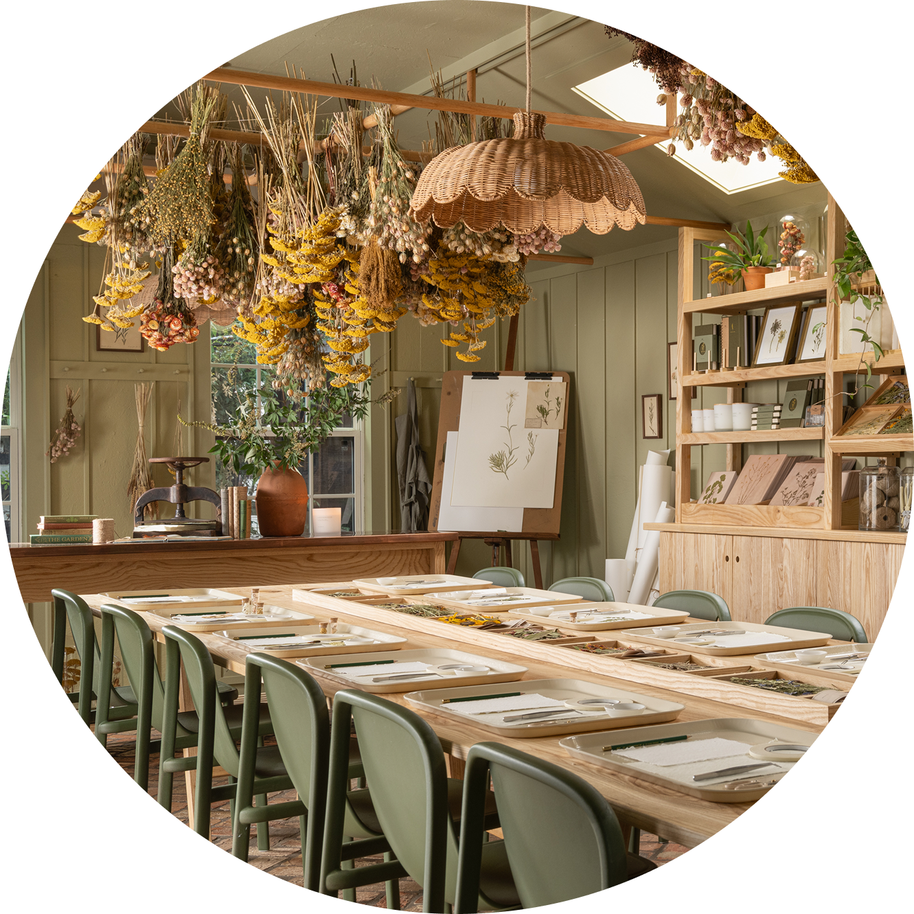 Long wooden table with green chairs in a room with hanging dried plants and bookshelves.  Table is set for a pressed floral workshop at the herbarium at the Silos in Waco.