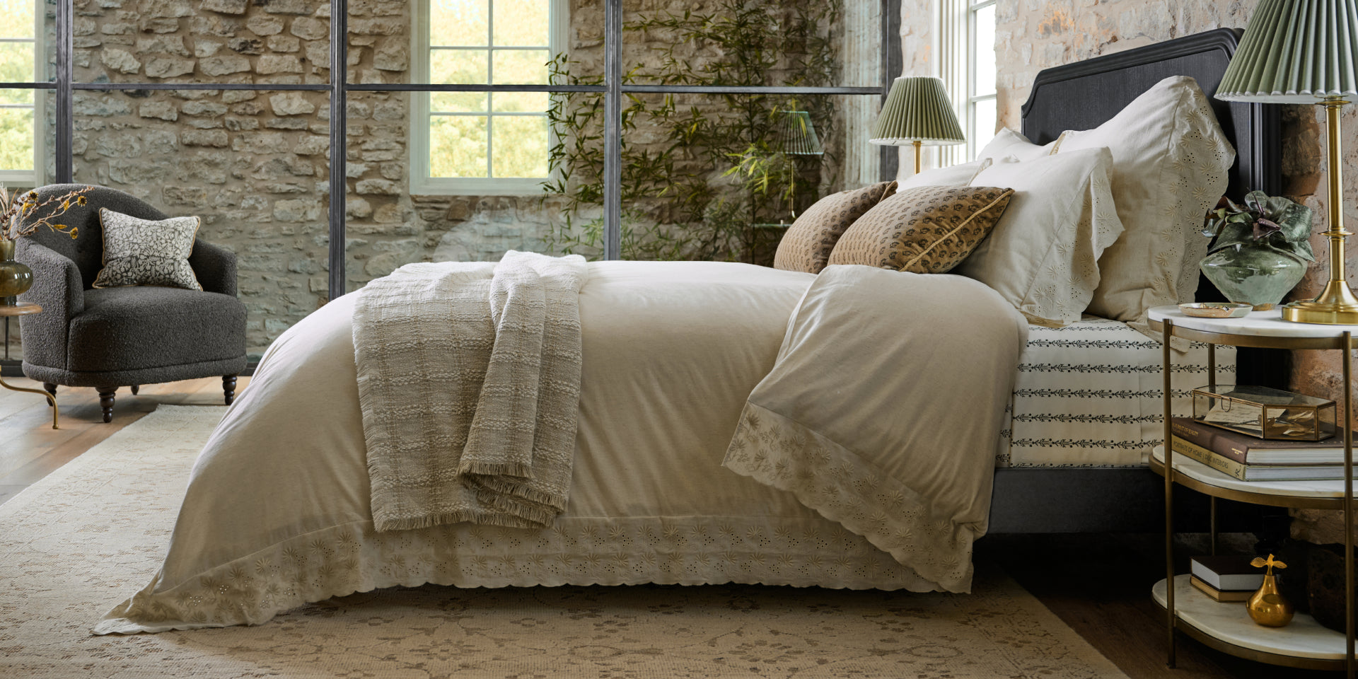 Neatly made bed in a room with stone walls and large windows.  Shop all Bedding.