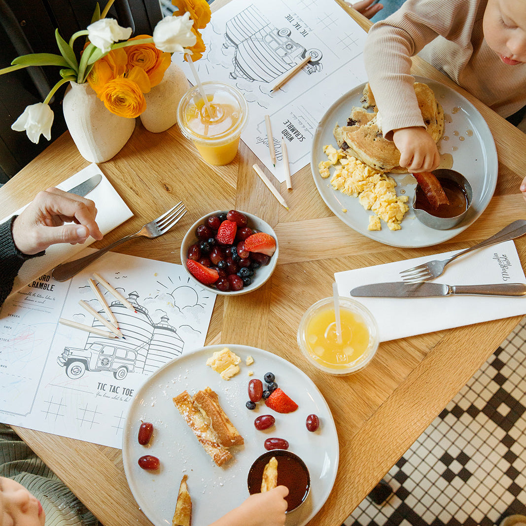 Dining table with plates of food, drinks, and coloring sheets with children around.