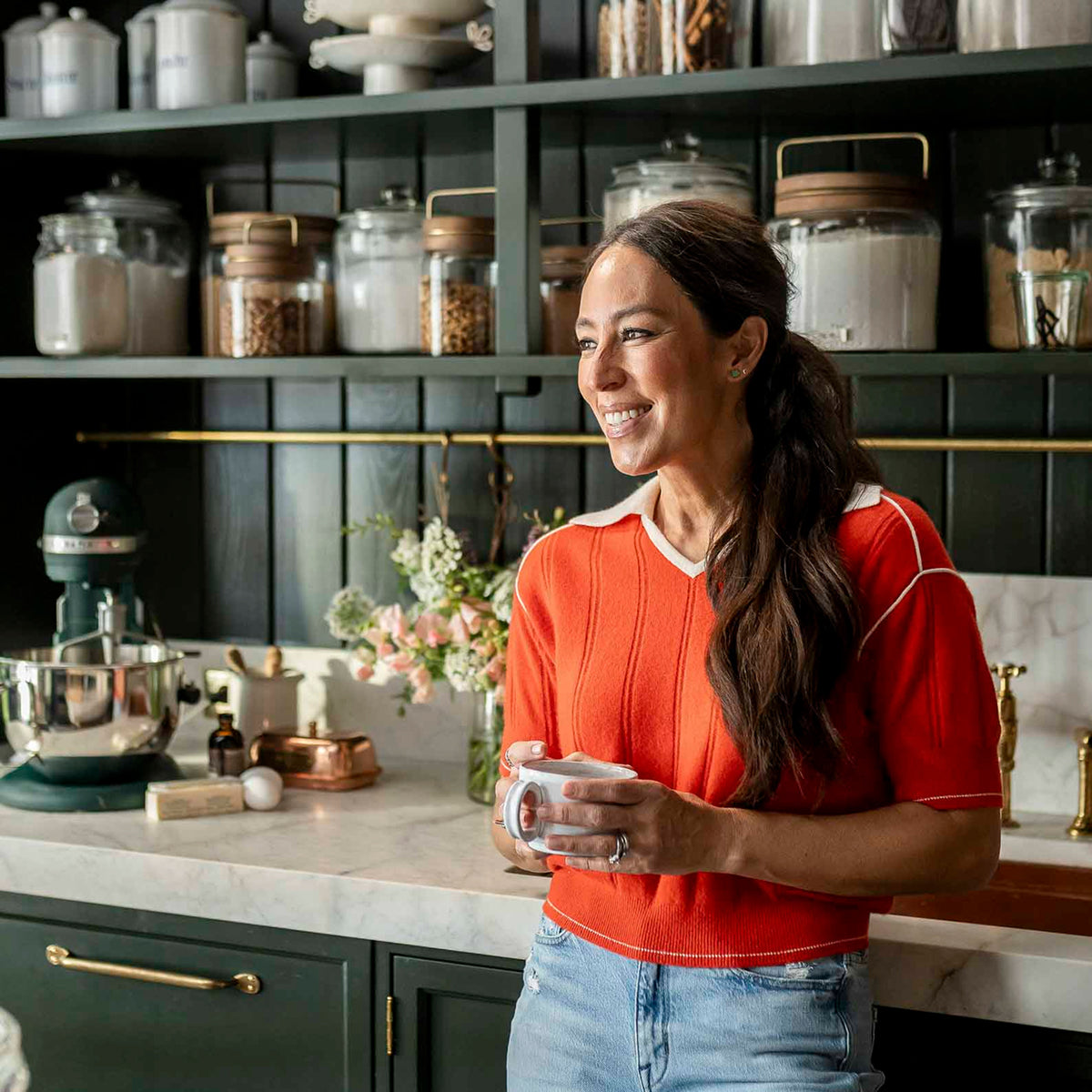 Joanna Gaines in her butler's pantry holding a mug, with shelves of canisters and a mixer in the background.