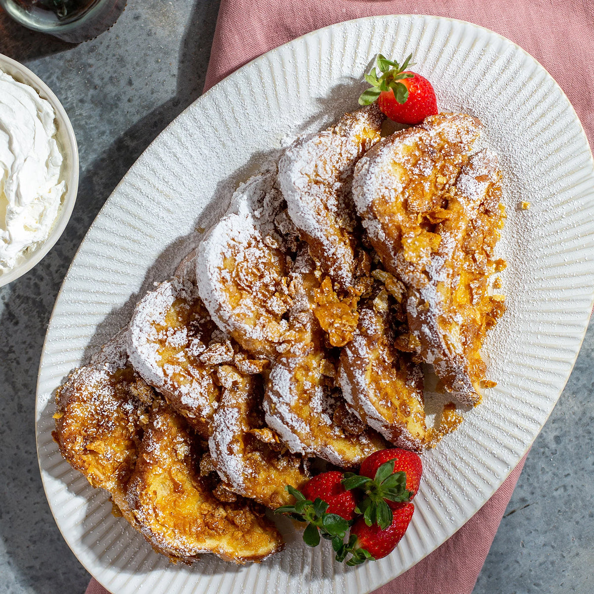 Crunch French Toast with powdered sugar on a white plate with strawberries, served with a side of whipped cream.