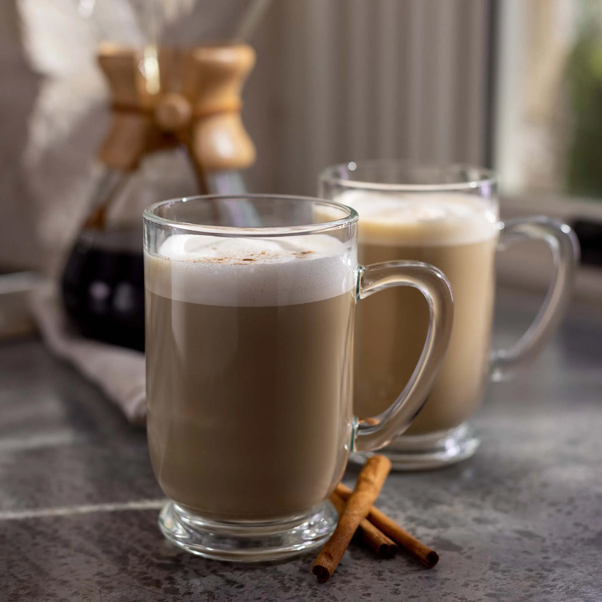 Two glass mugs of Snickerdoodle Coffee with whipped cream on a stone surface, with cinnamon sticks nearby.