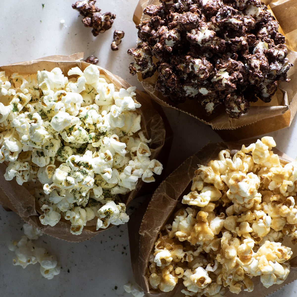 Three bowls of popcorn, two with white popcorn and one with chocolate-covered popcorn, on a textured surface.