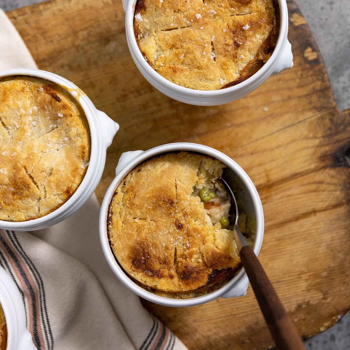Three small pot pies in white ramekins on a wooden board with a striped cloth.