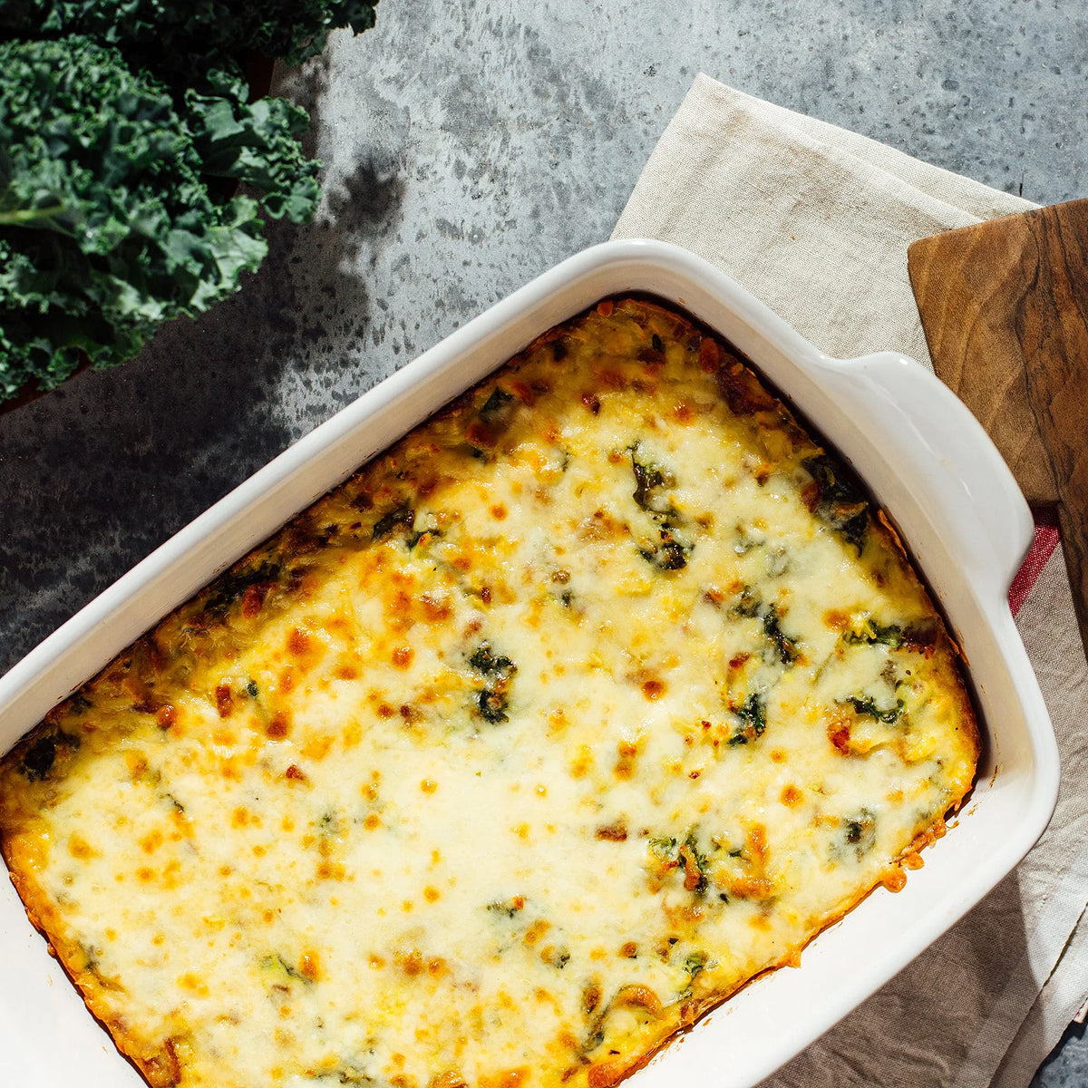 Cheesy Kale and Bacon Hash Brown Casserole in a white dish on a wooden surface.