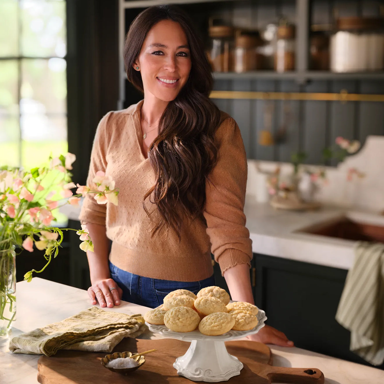 Joanna Gaines standing in her butler's pantry with Magnoliass Table Classic Sugar Cookies on a cake stad on her kitchen island.