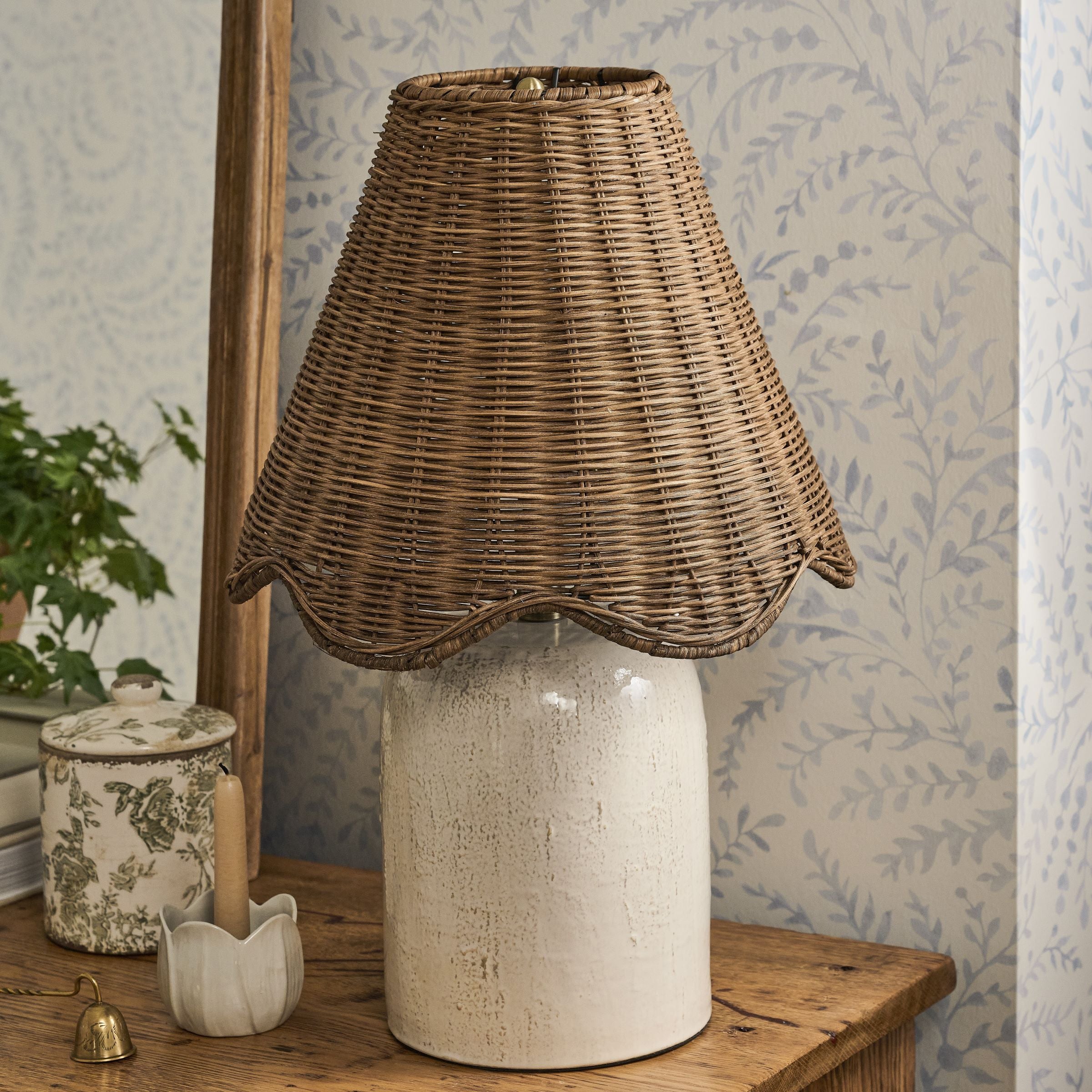 Decorative lamp with wicker shade on a wooden table against a floral wallpapered wall.