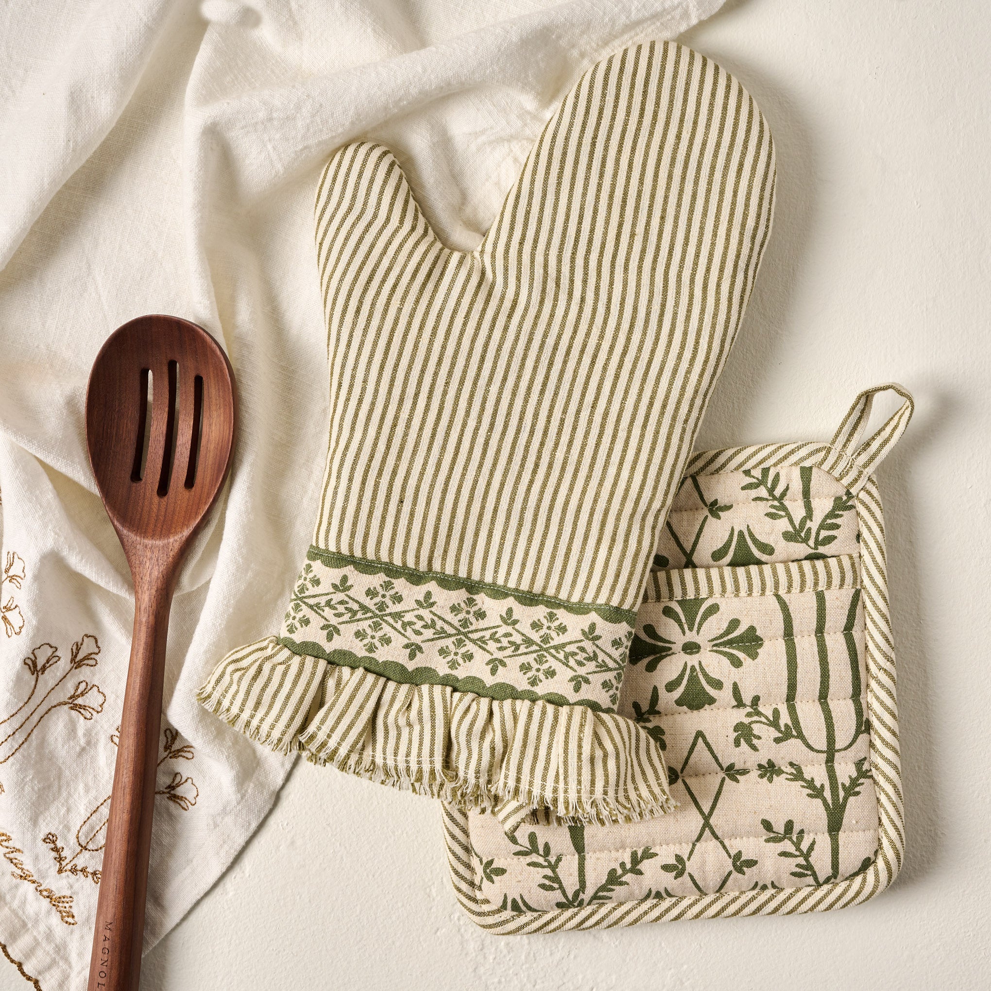 Magnoliass Olive Pin Stripe Oven Mitt + Pot Holder Set shown with wooden spoon and tea towel
