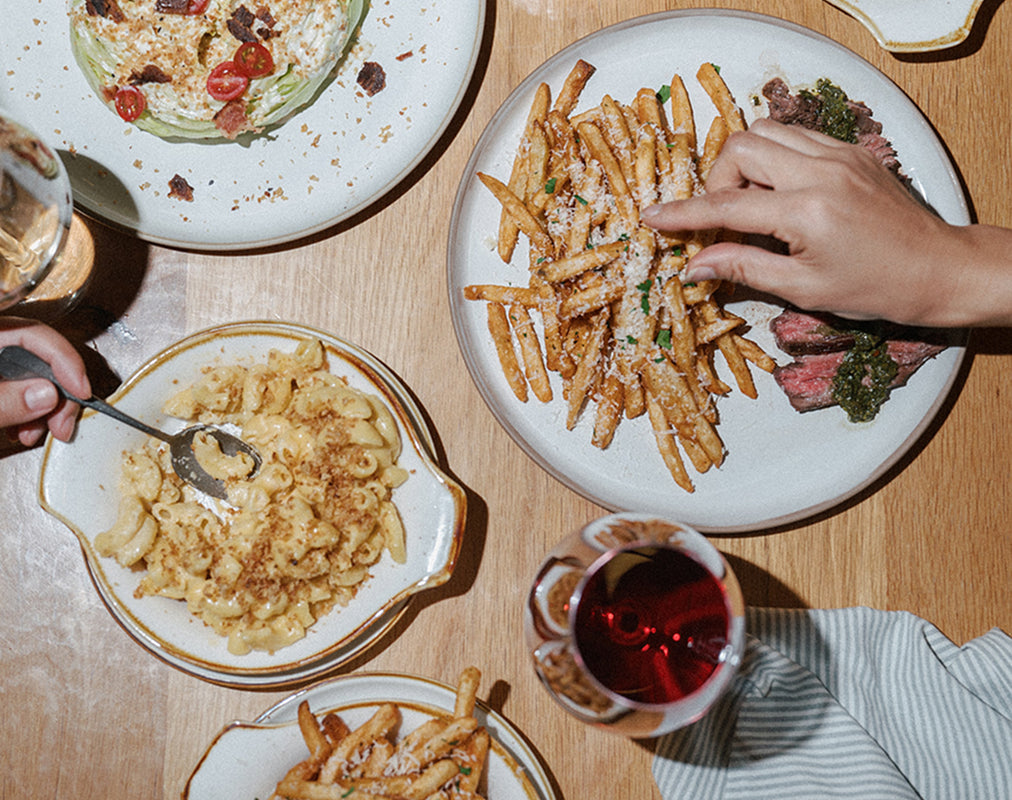 Diverse food dishes on a table with a hand reaching for fries. Book your holiday gathering.  Celebrate meaningful moments this season with an event at Magnoliass Table.  Inquire here.