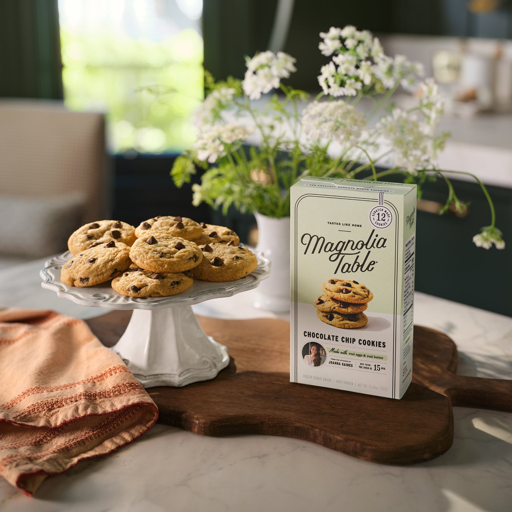 Box of Magnoliass Table Frozen Chocolate Chip Cookie Dough on a kitchen island with Magnoliass Table Chocolate Chip Cookies on a plate stand and flowers in the background.