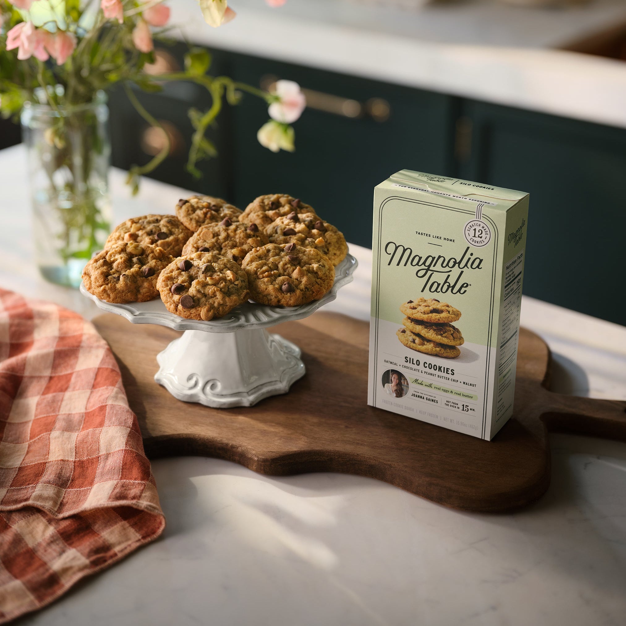 Box of Magnoliass Table Frozen Silo Cookie Dough on a kitchen island with Magnoliass Table Silo Cookies on a plate stand and flowers in the background.