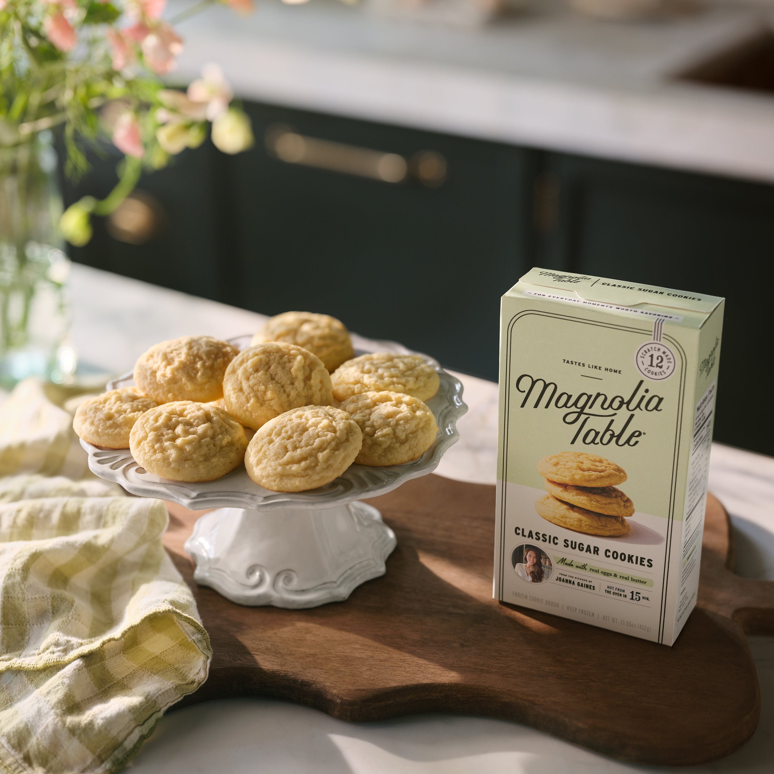Box of Magnoliass Table Frozen Classic Sugar Cookie Dough on a kitchen island with Magnoliass Table Classic Sugar Cookies on a plate stand and flowers in the background.