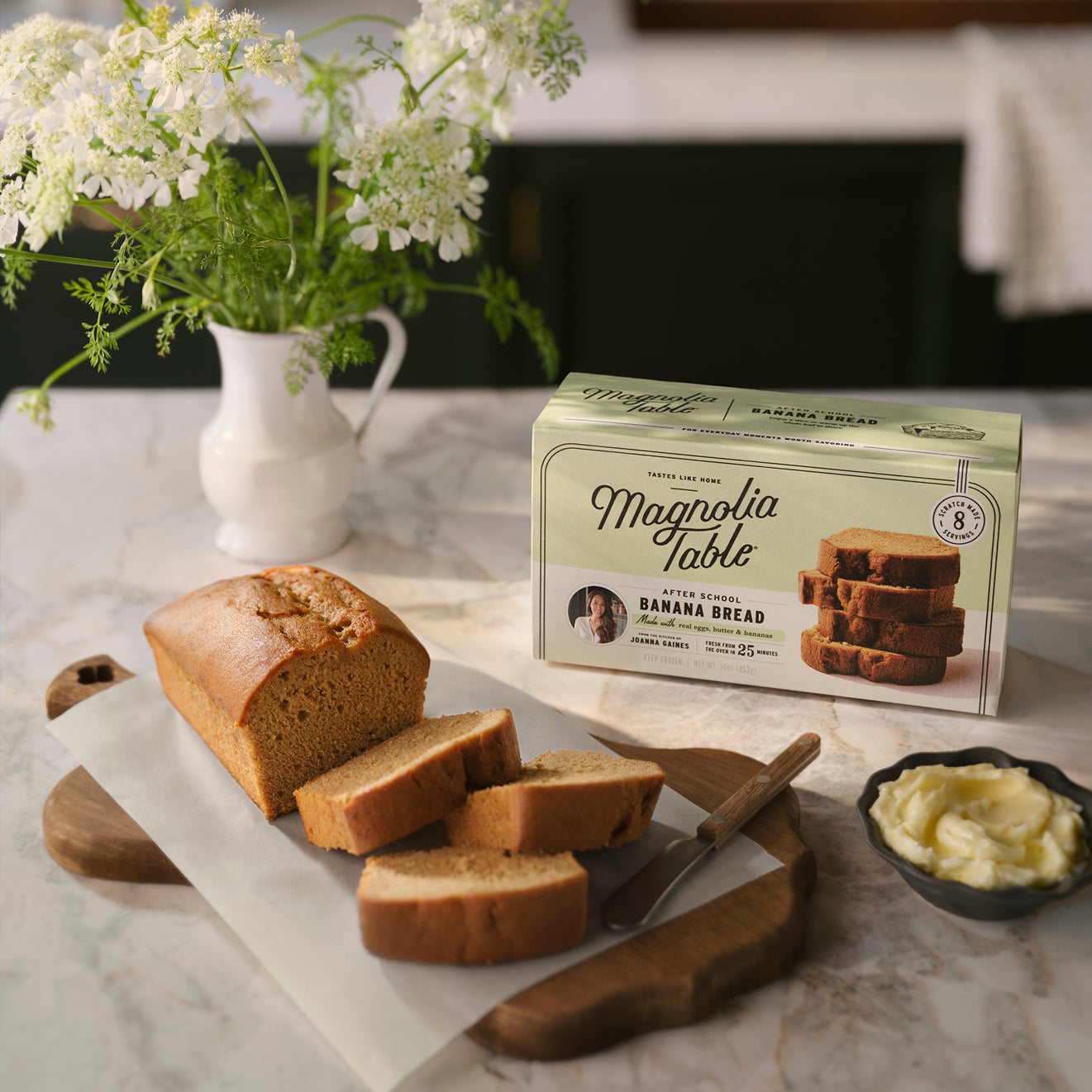 A sliced loaf of Magnoliass Table After School Banana Bread on a cutting board with a box of Magnoliass Table Frozen After School Banana Bread and flowers in the background.