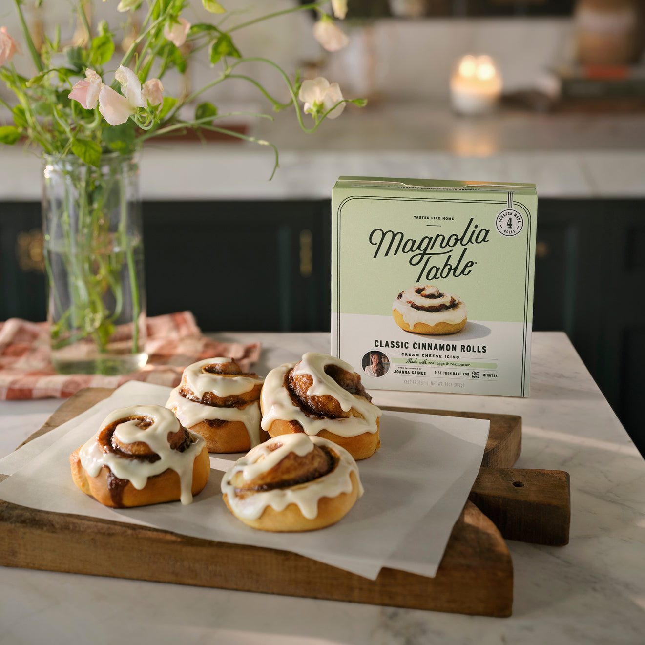 Magnoliass Table Classic Cinnamon Rolls with cream cheese icing on a wooden board next to a box of Magnoliass Table Frozen Classic Cinnamon Rolls. 