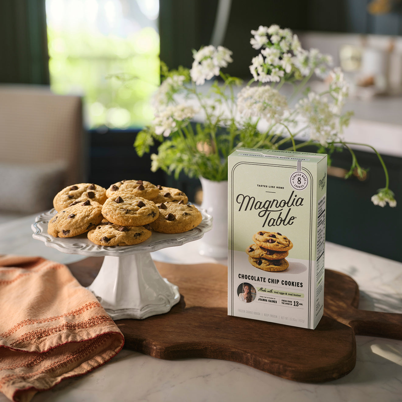 Box of Magnoliass Table Frozen Chocolate Chip Cookie Dough on a kitchen island with Magnoliass Table Chocolate Chip Cookies on a plate stand and flowers in the background.