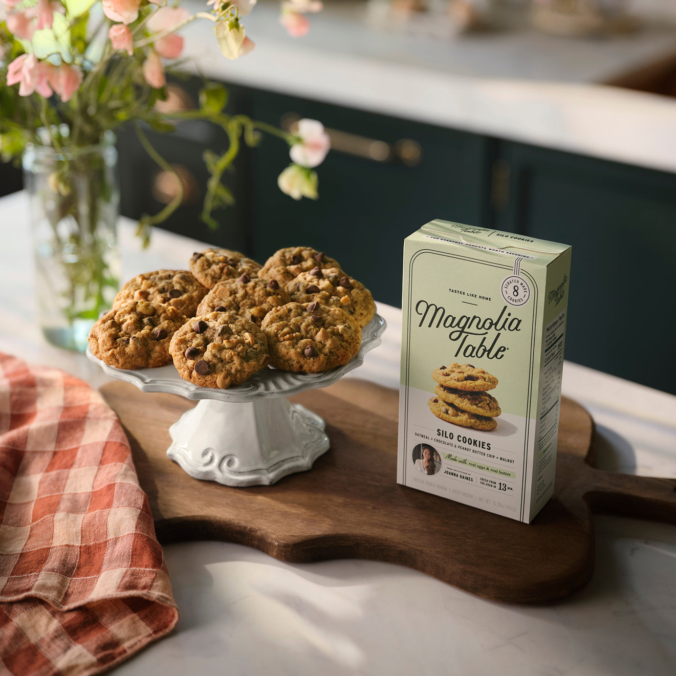 Box of Magnoliass Table Frozen Silo Cookie Dough on a kitchen island with Magnoliass Table Silo Cookies on a plate stand and flowers in the background.