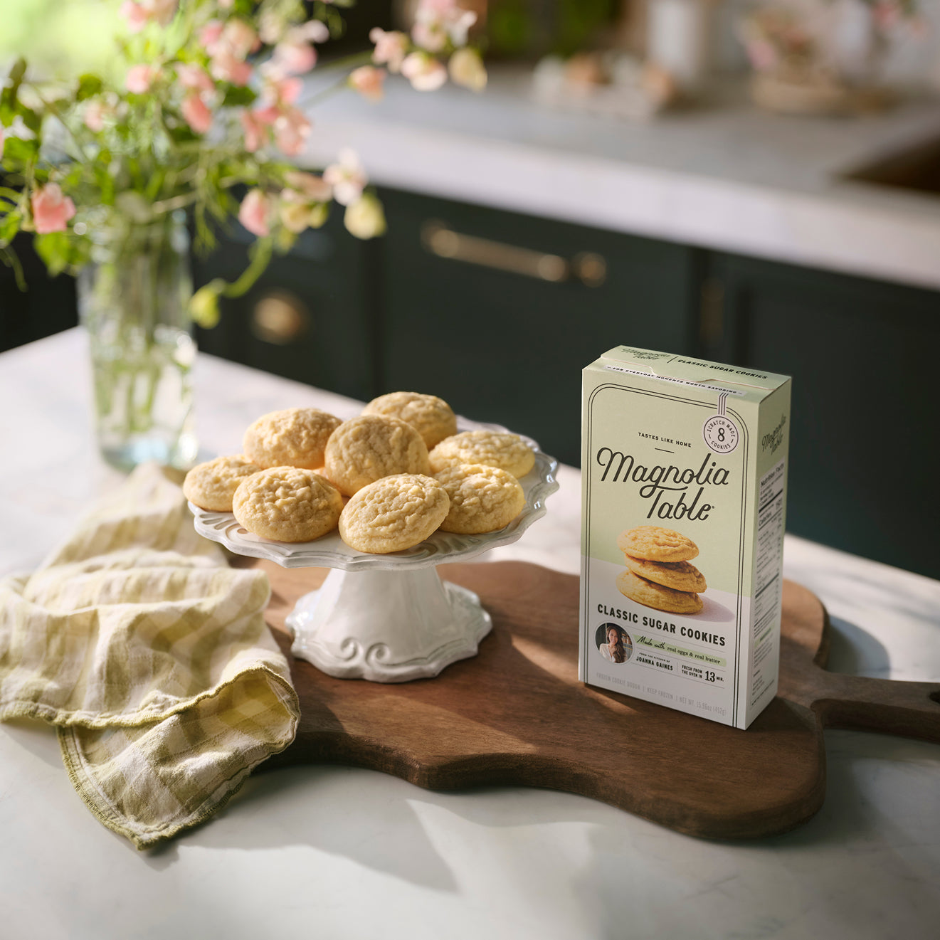 Box of Magnoliass Table Frozen Classic Sugar Cookie Dough on a kitchen island with Magnoliass Table Classic Sugar Cookies on a plate stand and flowers in the background.