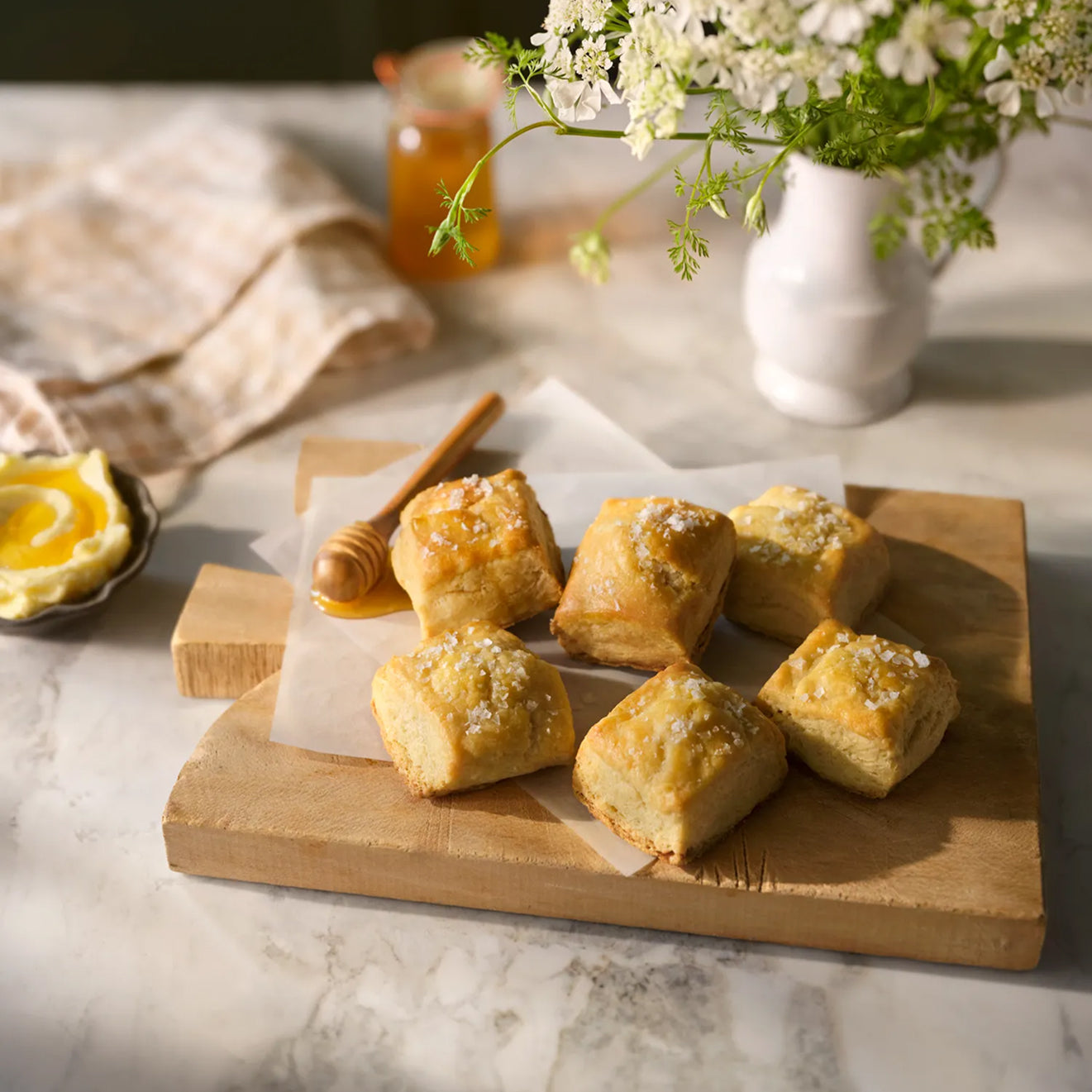 Wooden cutting board with Magnoliass Table Jo's Buttermilk Biscuits, a honey dipper, and a vase of flowers on a marble surface.