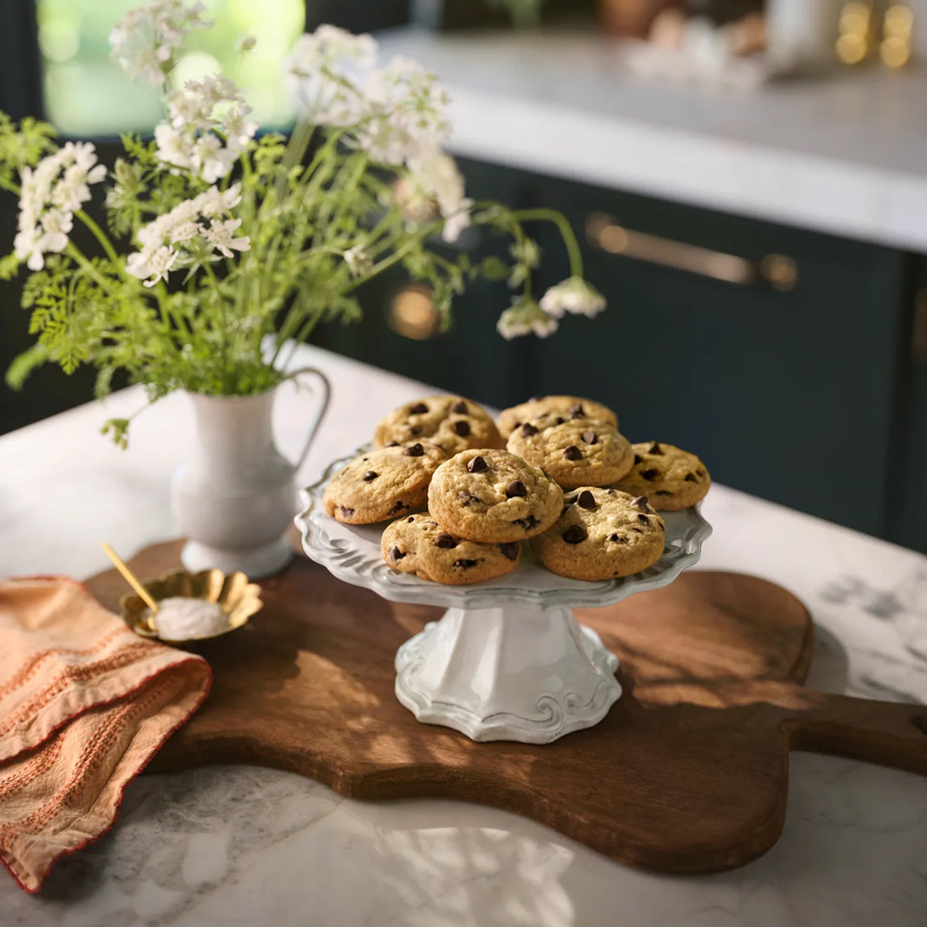 Magnoliass Table Chocolate Chip Cookies on a white cake stand with a vase of flowers and wooden cutting board in the background.