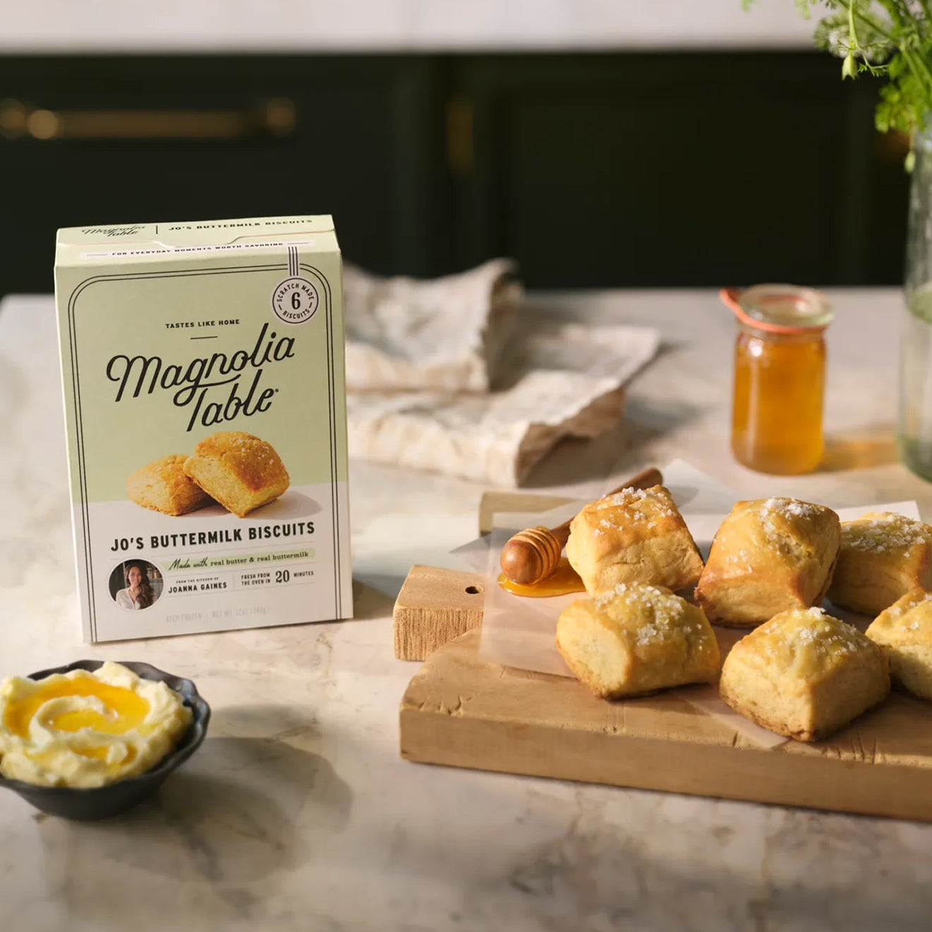 Box of Magnoliass Table Frozen Jo's Buttermilk Biscuits on a marble countertop with Magnoliass Table Jo's Buttermilk Biscuits and honey on a wooden cutting board.