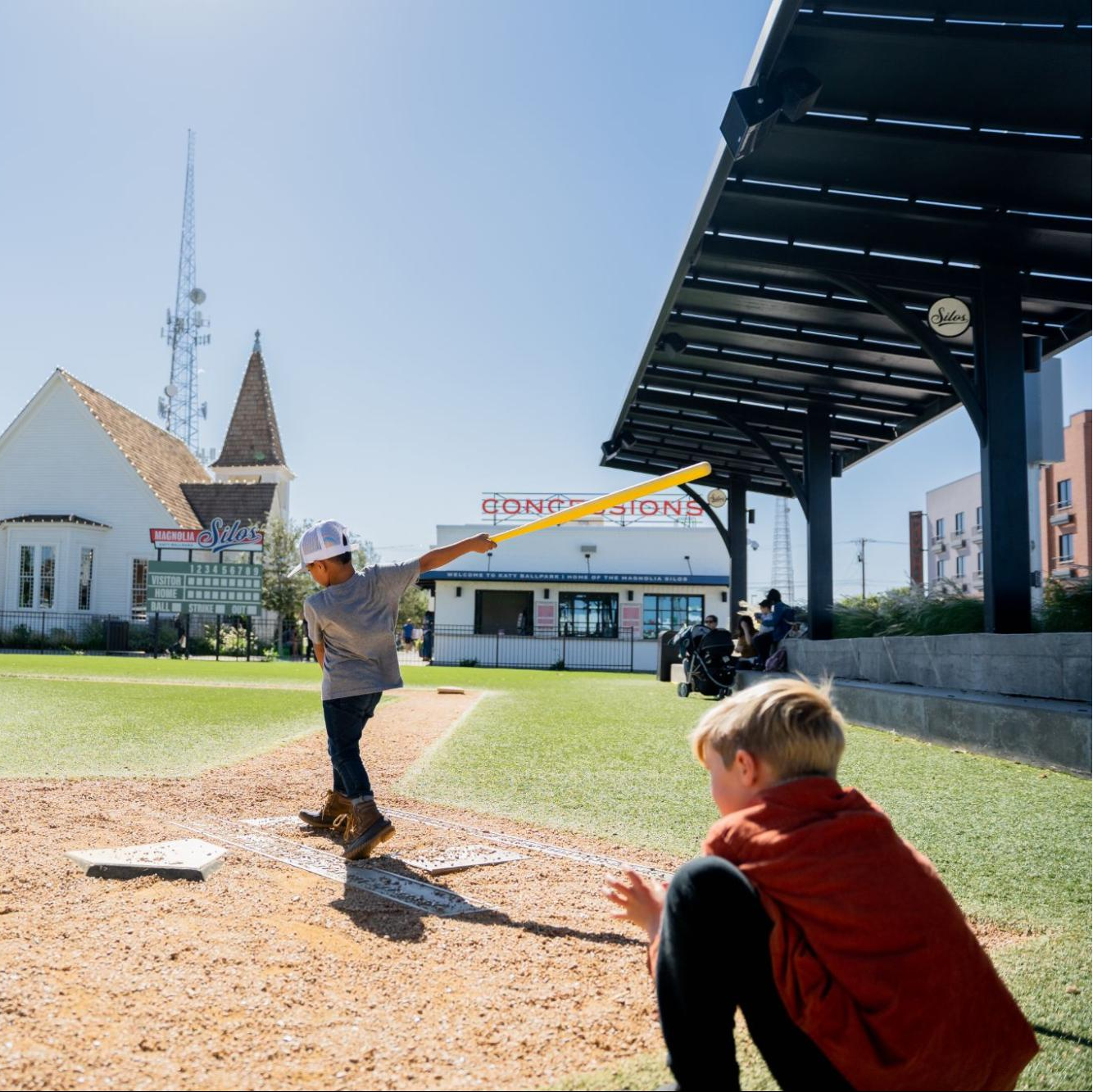 Two children playing baseball at Katy Ballpark on a sunny day with a clear blue sky.