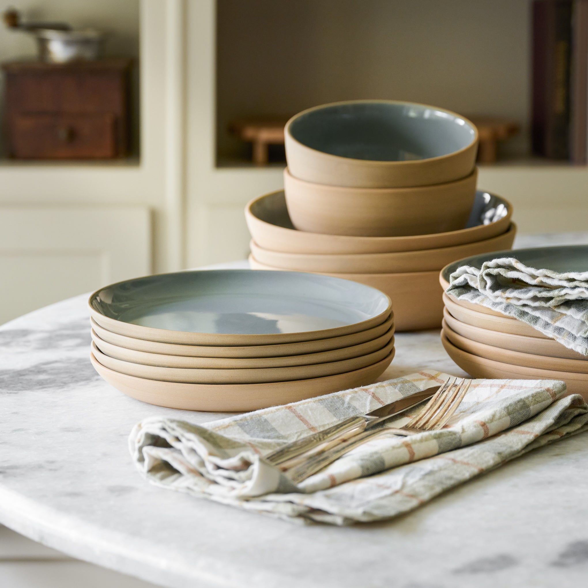 Lakelynn plates and bowls with blue glazed interior and tan exterior stacked on countertop next to plaid napkins and silverware