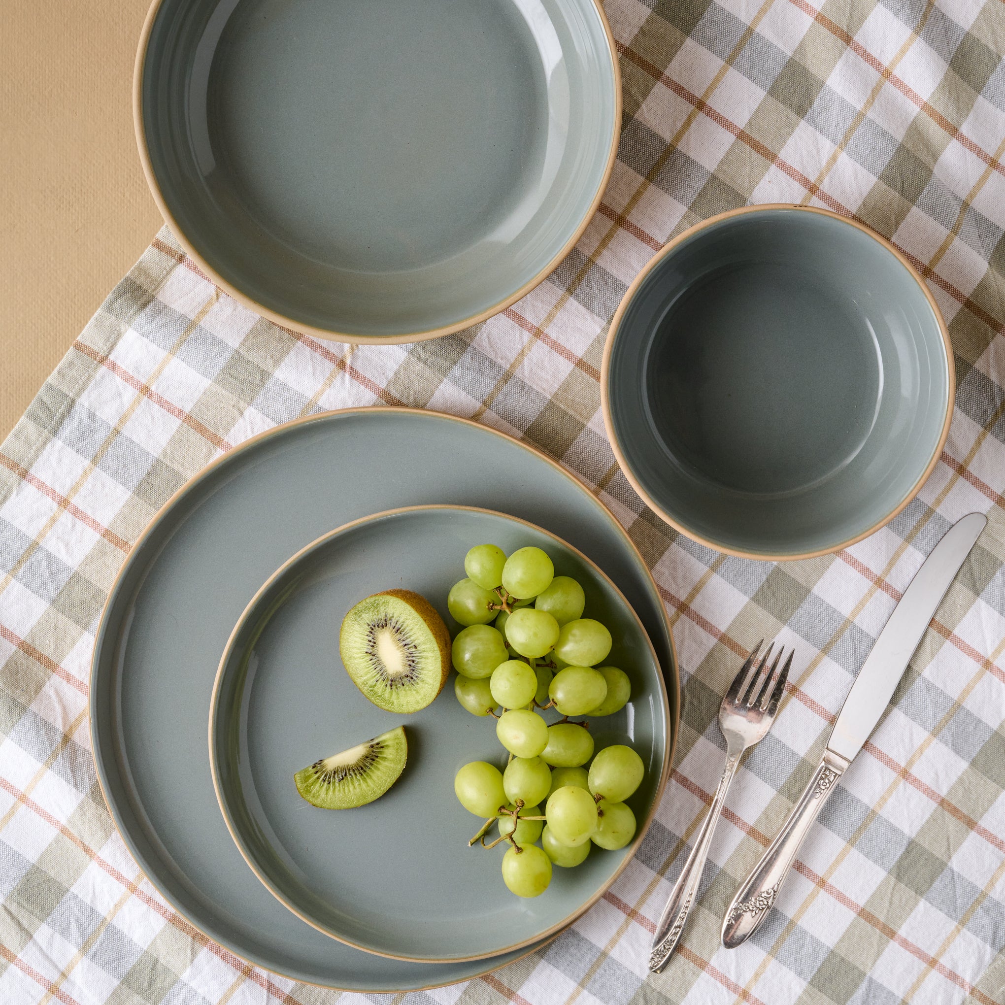 Lakelynn plates with a glazed blue gray interior and a tan exterior on tabletop with a plaid tablecloth. Grapes and kiwi are on the salad plate. 