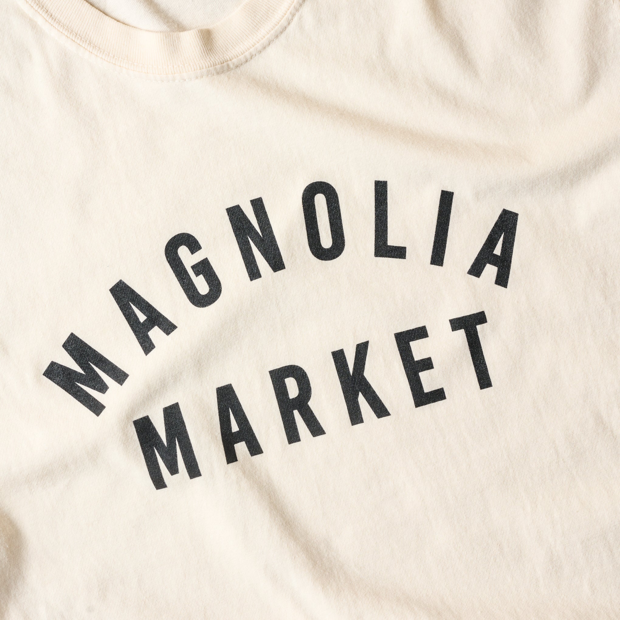 close up of design on the front of Magnoliass market ivory shirt that reads Magnoliass Market in plain black text. 