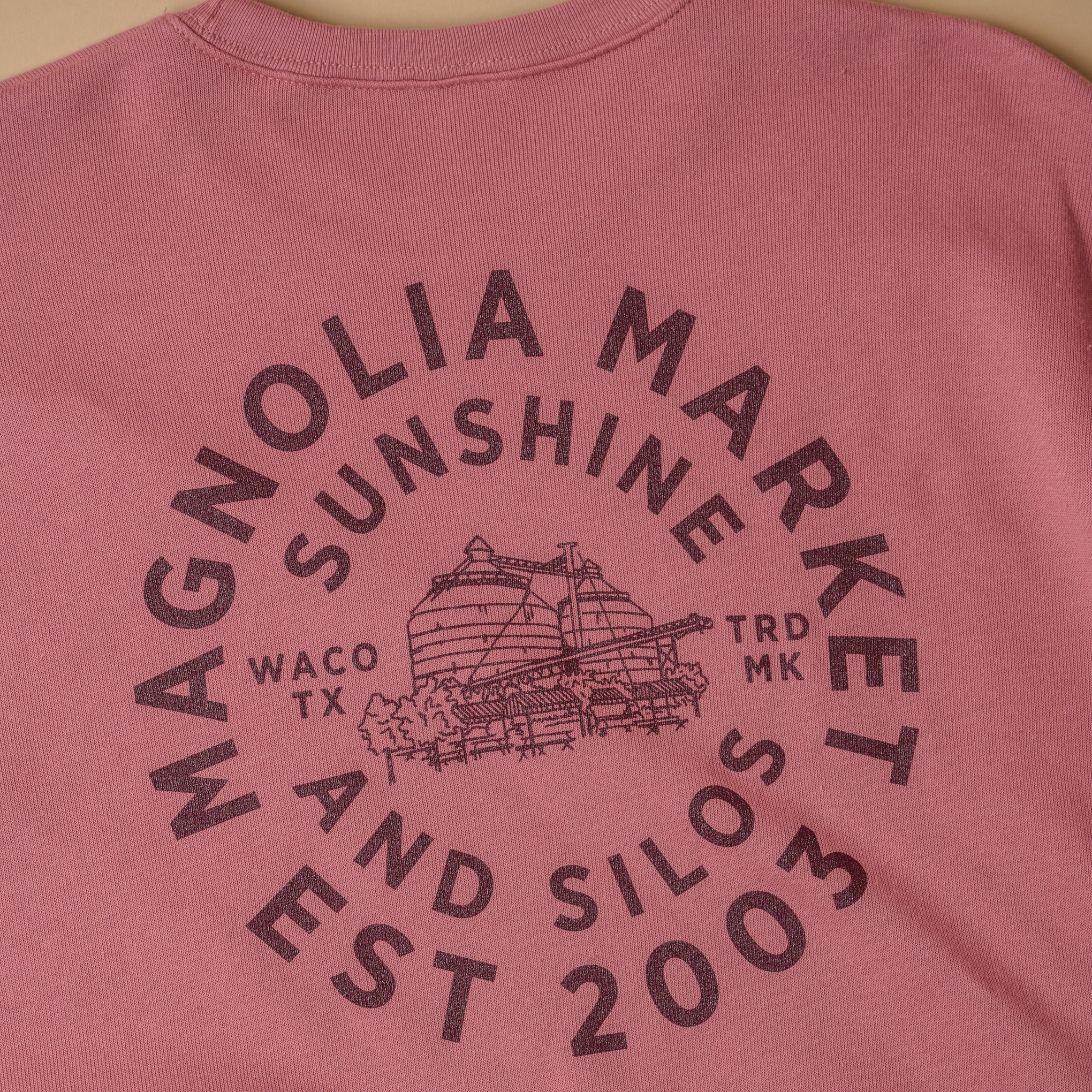 Close up of back of mauve colored sweatshirt in a circular shape reads Magnoliass Market Est 2003. There is an inner circle of text that reads sunshine and silos. In the center of the circle of text is a picture of the silos and on either side of the picture reads Waco TX and TRD MK. Sweatshirt on model with back turned. All text is in maroon color. 