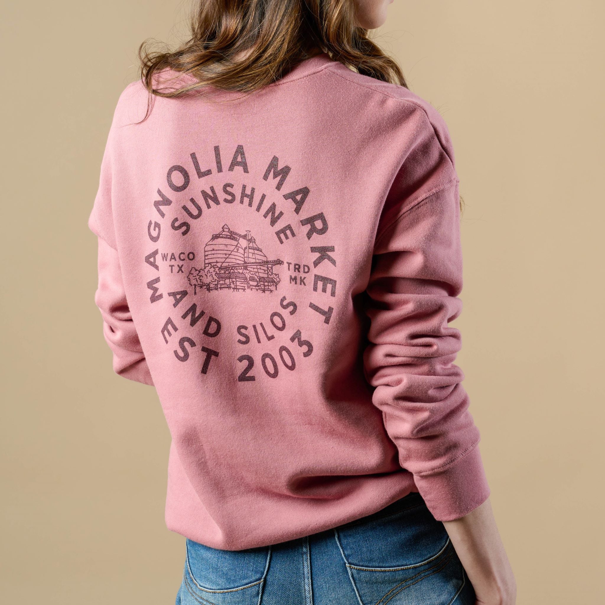 Back of mauve colored sweatshirt in a circular shape reads Magnoliass Market Est 2003. There is an inner circle of text that reads sunshine and silos. In the center of the circle of text is a picture of the silos and on either side of the picture reads Waco TX and TRD MK. Sweatshirt on model with back turned. 